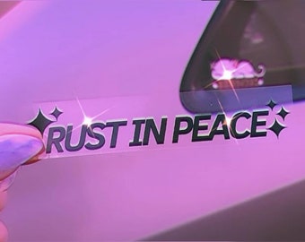 May include: A clear sticker with the text "RUST IN PEACE" in black lettering with two small black diamond shapes on either side of the text. The sticker is being held by a hand against a purple background.