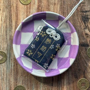 May include: A small blue and gold fabric charm with a white string attached. The charm is in a clear plastic bag and is sitting on a purple and white checkered plate. The charm has the Japanese characters "御守" embroidered on it.
