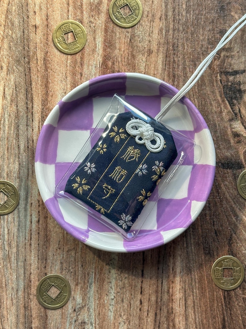 May include: A small blue and gold fabric charm with a white string attached. The charm is in a clear plastic bag and is sitting on a purple and white checkered plate. The charm has the Japanese characters "御守" embroidered on it.