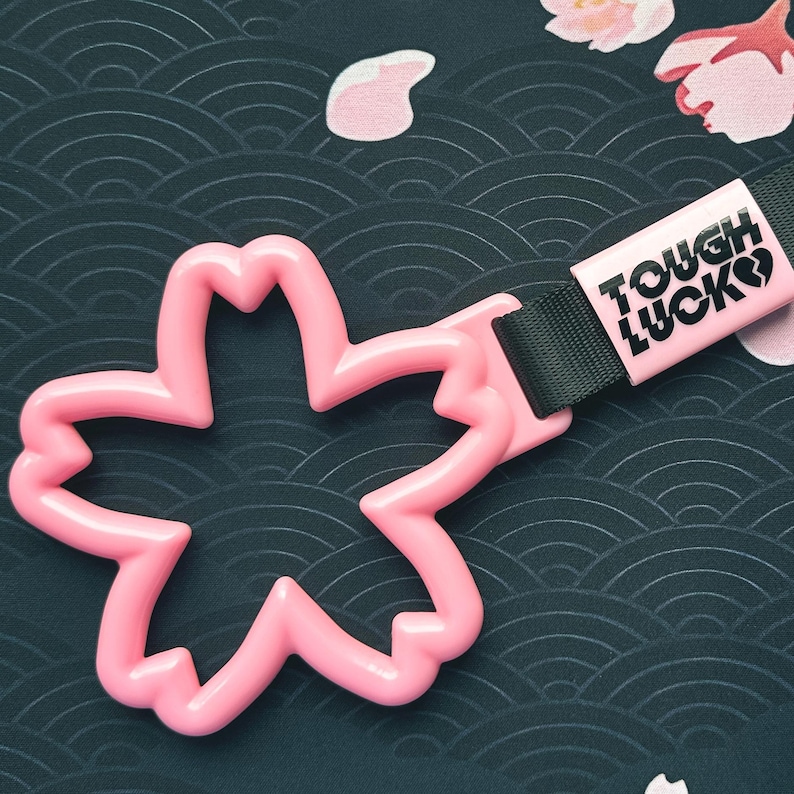 Może przedstawiać: R&oacute;żowy plastikowy brelok do samochodu w kształcie kwiatu wiśni z czarnym paskiem i r&oacute;żową metką z napisem "Tough Luck" czarnymi literami.