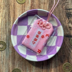May include: A pink Japanese good luck charm with white and red embroidery. The charm is on a purple and white checkered plate with a pink string attached. The charm has the Japanese characters "御守" embroidered on it.