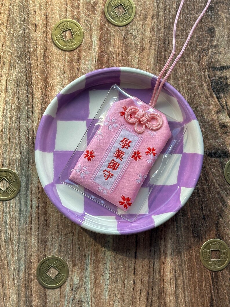 May include: A pink Japanese good luck charm with white and red embroidery. The charm is on a purple and white checkered plate with a pink string attached. The charm has the Japanese characters "御守" embroidered on it.