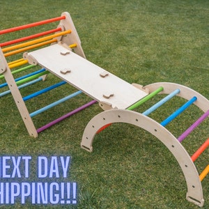 May include: Wooden climbing frame with a rainbow ladder and a slide. The frame is made of natural wood and has a light brown finish. The ladder has colorful rungs and the slide is a simple, flat surface. The frame is set up on a green grassy lawn.