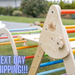 May include: Wooden climbing frame with rainbow colored rungs. The frame is made of natural wood and has a triangular shape. The frame is labeled "JR. CLIMBER".