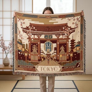 May include: A woven throw blanket featuring a detailed illustration of a Tokyo street scene with a large gate, lanterns, and pagodas. The blanket has a beige background with brown, red, and green accents. The word "TOKYO" is displayed prominently.