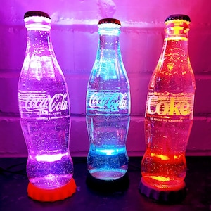 May include: Three glass Coca-Cola bottles with colorful LED lights inside. The bottles are filled with clear liquid and have a frosted glass effect. The bottles are sitting on a table with a brick wall background.