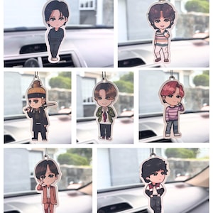 May include: A collection of seven hanging car air fresheners, each featuring a unique cartoon-style illustration of a person with distinct hair colors and outfits, including a warrior in traditional attire and various casual clothing styles.