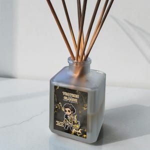 May include: A frosted glass reed diffuser with wooden reeds. The label reads "Teatime Claude reed diffuser" and features a cartoon character. The diffuser contains a liquid and is on a white surface.