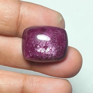 May include: A cushion-cut ruby gemstone with a deep magenta-red hue and a polished, smooth surface. The stone displays a natural, textured internal structure with visible inclusions and a bright white light reflection on its top surface.