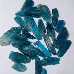 Op de afbeelding: Een verzameling ruwe, blauwe kyanietkristallen. De kristallen vari&euml;ren in grootte en vorm en vertonen een reeks blauwtinten, van licht turkoois tot diep indigo. De ruwe textuur en natuurlijke formaties zijn duidelijk zichtbaar.