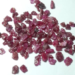 Puede incluir: Un conjunto de gemas de rubí en bruto y sin cortar. Los rubíes, de tamaños y formas variadas, presentan un color rojo intenso con algunas inclusiones más oscuras. Las piedras están dispersas sobre una superficie blanca, revelando su textura natural.