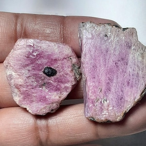 Könnte beinhalten: Zwei grob geschnittene, rosa Edelsteine mit kristalliner Struktur. Ein Stein hat einen dunklen Einschluss. Der andere Stein hat ein geschichtetes Aussehen. Die Steine werden in einer Hand gehalten und zeigen ihren natürlichen, unpolierten Zustand.