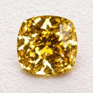 May include: A cushion-cut yellow gemstone with a brilliant faceted surface. The stone displays a vibrant, saturated golden-yellow hue with sharp geometric light reflections across its top surface, showcasing a high level of clarity and precision.