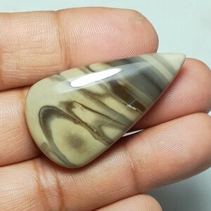 Könnte beinhalten: Ein polierter, tropfenförmiger Cabochon-Edelstein mit glatten, fließenden Bändern in den Farben Beige, Hellbraun und Dunkelbraun. Der Stein hat eine glänzende, reflektierende Oberfläche mit einem ausgeprägten Wirbelmuster, das natürlich wirkt.