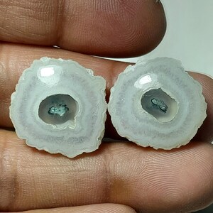 May include: Two polished, circular gemstones with a white and gray layered pattern. Each stone has a dark teal center and a rough, textured edge. The stones are held in a hand, showcasing their natural beauty.