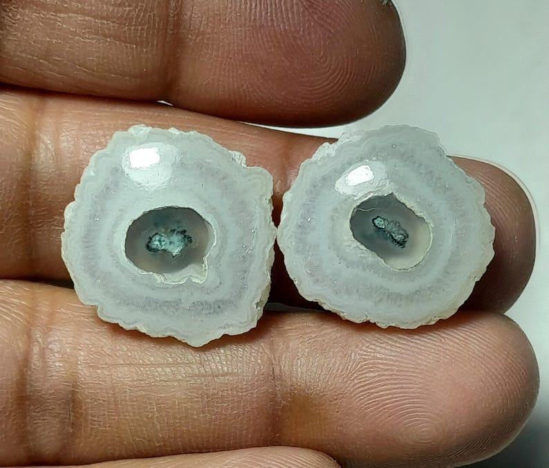 May include: Two polished, circular gemstones with a white and gray layered pattern. Each stone has a dark teal center and a rough, textured edge. The stones are held in a hand, showcasing their natural beauty.