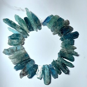 May include: A circular arrangement of raw, elongated kyanite crystal beads in varying shades of blue, teal, and grey. Each crystal features a natural, jagged texture and is strung together on a thin, white cord, creating a continuous loop.