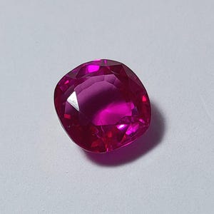 May include: A faceted, cushion-cut ruby gemstone. The gem is a vibrant, deep pink color with multiple facets that reflect light. The gemstone is set against a white background.