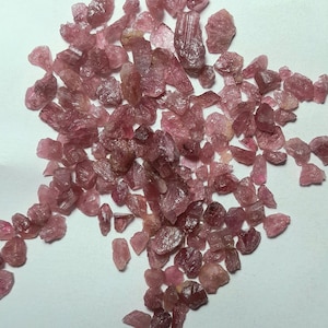 May include: A collection of rough, pink tourmaline gemstones. The gemstones vary in size and shape, with a range of pink hues from light to deep rose. The stones are scattered on a white surface, showcasing their natural, unpolished texture.