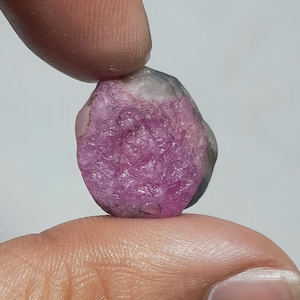 May include: A raw, uncut gemstone featuring a vibrant, saturated pink centre that transitions into a translucent, greyish-white outer edge. The stone has a rough, crystalline texture with visible facets and natural mineral inclusions throughout.