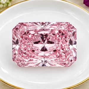 Pode incluir: Uma grande pedra preciosa rosa de corte retangular radiante com uma superfície facetada que reflete a luz num padrão geométrico. A pedra está posicionada centralmente num pequeno prato redondo de cerâmica branca com uma borda dourada.