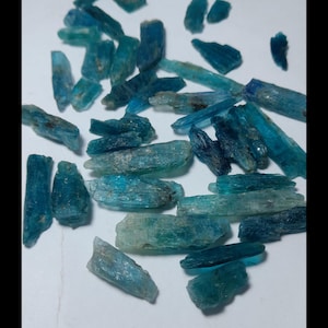 Op de afbeelding: Een verzameling ruwe kyanietkristallen in tinten blauw en teal. De kristallen zijn langwerpig en hebben een natuurlijke, ruwe textuur. Ze zijn verspreid op een wit oppervlak, waarbij hun verschillende vormen en maten worden getoond. Deze edelstenen worden vaak gebruikt in sieraden.