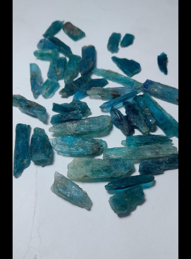 Op de afbeelding: Een verzameling ruwe kyanietkristallen in tinten blauw en teal. De kristallen zijn langwerpig en hebben een natuurlijke, ruwe textuur. Ze zijn verspreid op een wit oppervlak, waarbij hun verschillende vormen en maten worden getoond. Deze edelstenen worden vaak gebruikt in sieraden.