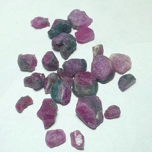 May include: A collection of raw, unpolished ruby gemstones in various sizes and shapes. The stones exhibit a mix of deep magenta, vibrant pink, and dark green hues with natural, irregular textures and opaque to translucent crystalline surfaces.