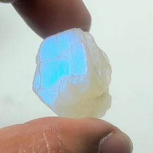 May include: A rough, uncut moonstone gemstone held between fingers. The stone has a creamy white base color with a flash of blue iridescence. The surface shows natural cracks and imperfections, highlighting its raw, organic form.