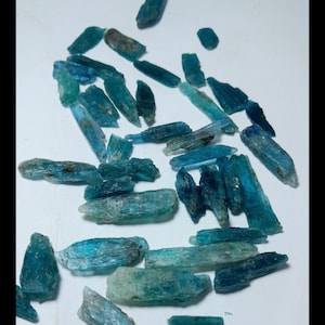 Op de afbeelding: Een verzameling ruwe kyanietkristallen in tinten blauw en teal. De kristallen vari&euml;ren in grootte en vorm, sommige zijn langwerpig en andere dikker. De natuurlijke stenen zijn verspreid op een witte ondergrond, waarbij hun unieke texturen en kleuren worden getoond.