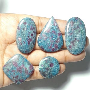 May include: Five polished gemstones in various shapes: oval, teardrop, and round. The stones have a teal base color with scattered red inclusions. The gemstones are held in a hand, showcasing their polished surfaces and unique patterns.