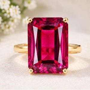 Könnte beinhalten: Ein Goldring mit einem großen, rechteckigen, smaragdgeschliffenen, tiefrosa Edelstein, der mit vier Krappen gefasst ist. Die Ringschiene ist aus glattem, poliertem Gold und bildet einen klaren Kontrast zu dem leuchtenden, facettierten Stein.