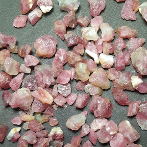 May include: A collection of raw, uncut pink tourmaline gemstones. The stones vary in size and shape, with a range of pink hues from light to deep rose. Some stones have white or clear inclusions, and all have a rough, natural texture.