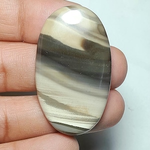 Könnte beinhalten: Ein ovaler, polierter Edelstein-Cabochon mit glatten, fließenden Bändern in Creme, Beige und Dunkelanthrazit. Der Stein hat eine hochglänzende Oberfläche mit reflektierenden Eigenschaften, die die natürlichen Schichtmuster des Steins hervorhebt.