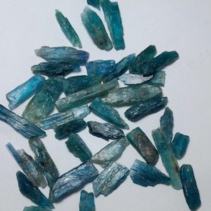 Op de afbeelding: Een verzameling ruwe kyanietkristallen in tinten blauw en teal. De kristallen vari&euml;ren in grootte en vorm, met een ruwe, getextureerde uitstraling. De kyanietstukken zijn verspreid op een witte ondergrond.
