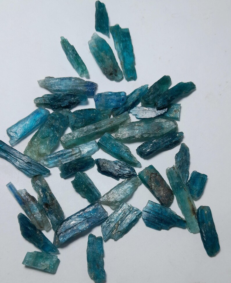 Op de afbeelding: Een verzameling ruwe kyanietkristallen in tinten blauw en teal. De kristallen vari&euml;ren in grootte en vorm, met een ruwe, getextureerde uitstraling. De kyanietstukken zijn verspreid op een witte ondergrond.