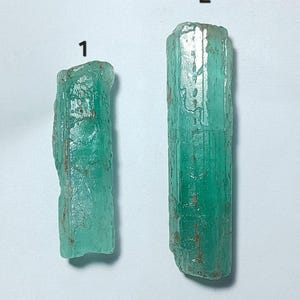 Op de afbeelding: Twee ruwe, ongesneden smaragdkristallen. De kristallen hebben een levendige groene kleur met ruwe, getextureerde oppervlakken. De eerste kristal is korter, terwijl de tweede langer is. Beide hebben bruine insluitsels. De achtergrond is wit.