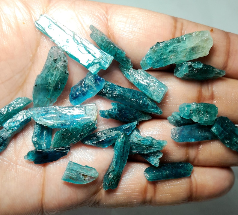 Op de afbeelding: Een verzameling ruwe, teal-kleurige kyanietkristallen. De kristallen vari&euml;ren in grootte en vorm, met een doorschijnende kwaliteit. De kyanietstukken worden in een hand gehouden, waardoor hun natuurlijke, ongepolijste staat wordt benadrukt. De kleur varieert van licht tot diep teal.