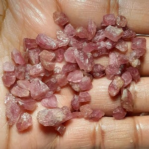 May include: A collection of small, rough-cut pink gemstones. The stones vary in size and shape, with a range of pink hues from light to deep rose. The gemstones are held in a hand, showcasing their natural, unpolished state. Ideal for jewelry making or collecting.