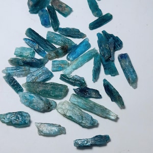 Op de afbeelding: Een verzameling ruwe, blauwe kyanietkristallen verspreid over een wit oppervlak. De kristallen vari&euml;ren in grootte en vorm en vertonen tinten van blauwgroen, turkoois en diepblauw. De ruwe textuur en natuurlijke formaties zijn zichtbaar.