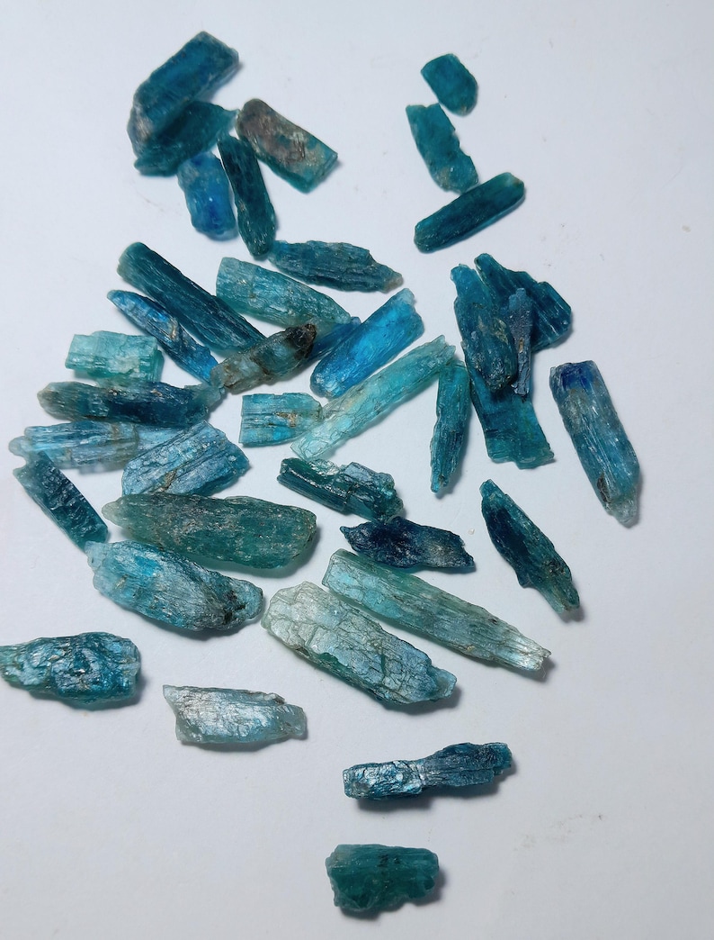 Op de afbeelding: Een verzameling ruwe, blauwe kyanietkristallen verspreid over een wit oppervlak. De kristallen vari&euml;ren in grootte en vorm en vertonen tinten van blauwgroen, turkoois en diepblauw. De ruwe textuur en natuurlijke formaties zijn zichtbaar.