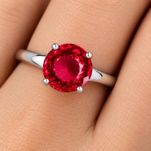 Puede incluir: Un anillo de tono plateado con una gran piedra preciosa roja vibrante, redonda y facetada, sujeta por un engaste de cuatro garras. La banda es lisa y pulida, estrechándose ligeramente a medida que se aleja del engaste central.