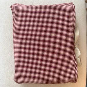 May include: A rectangular pillow with a red and white gingham pattern. The pillow is tied with a cream-colored ribbon on one side. The pillow is resting on a cream-colored surface.