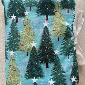 May include: A rectangular fabric item featuring a Christmas tree design. The trees are green and light green with white star toppers and colourful baubles, set against a light blue backdrop. A white ribbon is attached to the side.