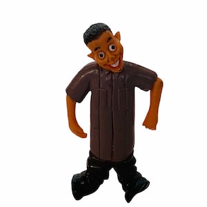 May include: A brown plastic figurine of a person with a wide smile and exaggerated features. The figurine is wearing a brown shirt and black trousers.