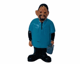 May include: A small figurine of a man in a blue shirt, black trousers, and black boots, holding a grey toolbox. The figurine has a dark beard and is standing upright.