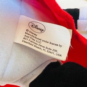 May include: A white tag with black text that reads "&copy; Disney Manufactured under license by Just Play, LLC 401 Fairway Drive Suite 200, Deerfield Beach, FL 33441, USA". The tag is attached to a red and white plush toy.