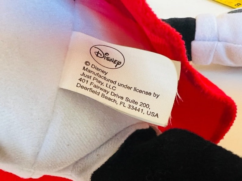 May include: A white tag with black text that reads "&copy; Disney Manufactured under license by Just Play, LLC 401 Fairway Drive Suite 200, Deerfield Beach, FL 33441, USA". The tag is attached to a red and white plush toy.