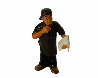 May include: A brown and black figurine of a person wearing a black baseball cap, a black t-shirt, blue jeans, and brown shoes. The person is holding a white notebook in their right hand and a pen in their left hand.