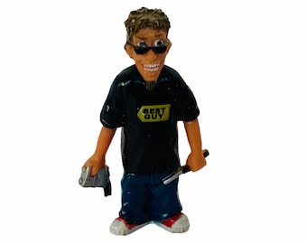 May include: A small plastic figurine of a person wearing a black t-shirt with the words "Best Buy" on it, blue jeans, and sunglasses. The figurine is holding a silver object in one hand and a black object in the other.